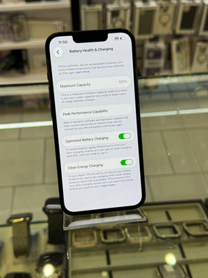 A smartphone displayed on a stand in a glass retail case, showing the iPhone Battery Health & Charging settings screen on its home screen.