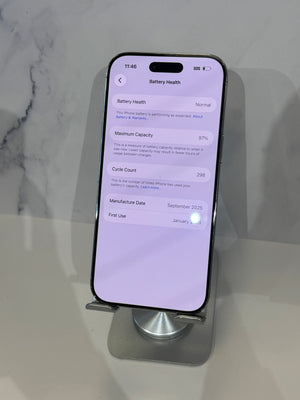 A silver iPhone displayed on a metallic stand, showing the battery health settings screen on its lit screen, set against a light marble-patterned background.