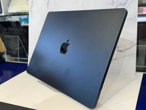 A dark blue-gray Apple laptop skin or protective case with a black Apple logo, shown standing on a white display stand in a store setting.