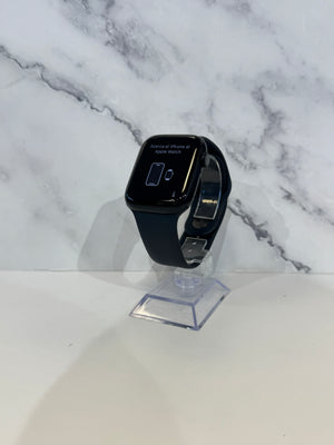 A black smartwatch with a rectangular face and black band, displayed upright on a clear acrylic stand against a white marble-patterned background.
