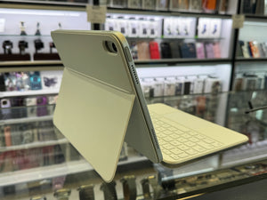 A light beige tablet case with an attached keyboard and kickstand, shown standing open on a glass display counter inside a retail store.