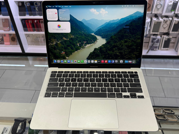 A silver Apple MacBook laptop with a black keyboard and trackpad, displaying a mountain river landscape on its screen, set on a glass display counter in a retail store.