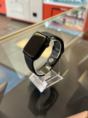 A black smartwatch with a rectangular face and black band, resting on a reflective dark glass surface with a small retail tag attached.
