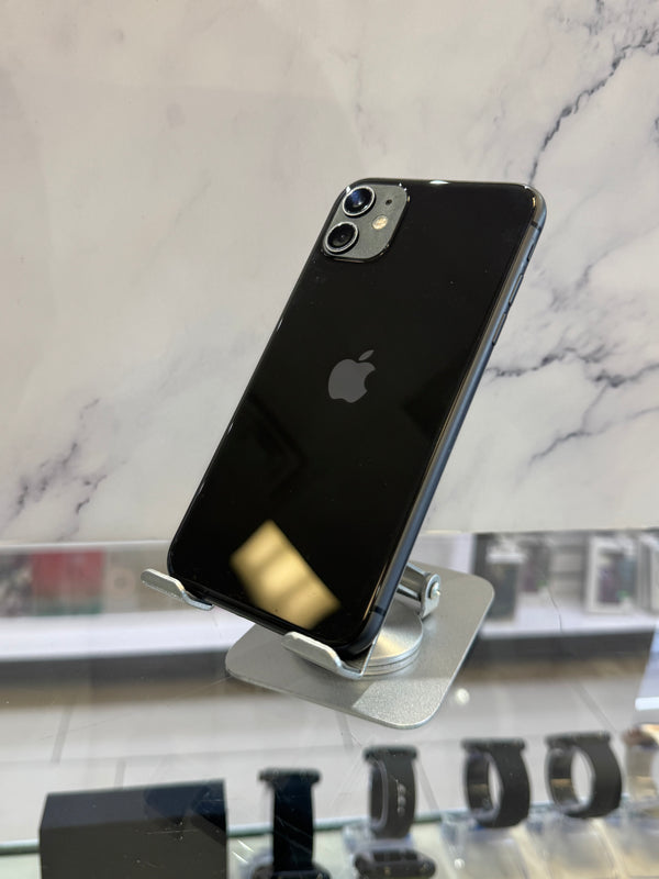 A black Apple iPhone with a glossy back is displayed on a silver metal stand inside a retail store, with a marble-patterned wall in the background.