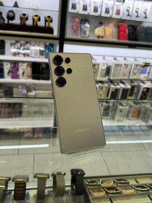 A silver Samsung smartphone with a three lens camera system displayed in a store setting with a blurred background