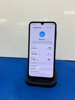 A black smartphone displayed upright on a black charging stand, showing the Android device care screen. The phone is photographed against a white wall with a blue surface below it.