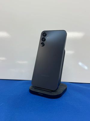 A Samsung smartphone in a dark gray or black finish, standing upright on a small black display stand against a white wall with a blue surface below.