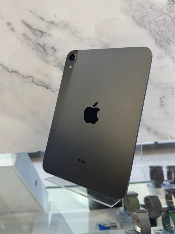 A dark gray Apple iPad shown from the back, standing on a clear display stand in front of a white marble-patterned background.