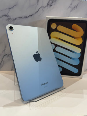 An iPad mini in light blue is displayed on a clear stand, with its retail box standing behind it against a white marble-patterned background.
