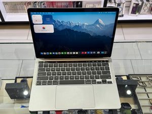 An open silver Apple MacBook laptop with a black keyboard and trackpad, displaying a mountain landscape desktop on a glass display counter in a retail store.