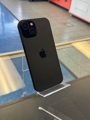 A black Apple iPhone shown from the back, resting on a clear acrylic stand on a glass display counter in a retail environment.
