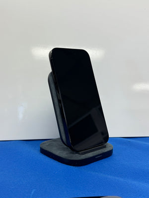 A black smartphone displayed upright on a dark gray charging stand against a white background with a blue surface below.