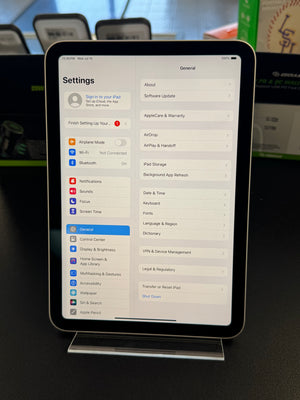 An iPad displaying the Settings menu on a clear stand, photographed in a retail or office environment with other devices and equipment blurred in the background.
