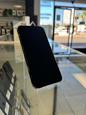 A black smartphone with a blank screen stands on a clear acrylic display stand inside a retail store. The background shows store fixtures, glass display cases, and a bright window or storefront with outdoor light.