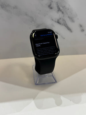 A black smartwatch with a black band displayed on a clear stand, showing a battery screen on the display. The watch is set against a marble-patterned background on a light surface.