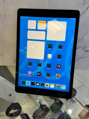 A black Apple iPad tablet standing upright on a clear display stand, showing the home screen with various app icons against a blue background. The tablet is photographed in a retail or showroom setting with a marble-patterned wall behind it and other accessories visible on a glass counter below.