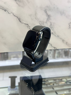 A black smartwatch with a dark gray mesh band displayed on a black stand, set against a white marble-patterned background.