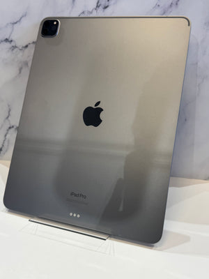 An Apple iPad Pro shown from the back, featuring a metallic dark gray finish and the black Apple logo in the center, resting on a white surface against a marble-patterned background.