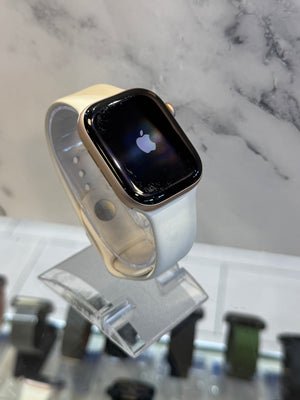 A smartwatch with a gold-toned case and a light beige band, displayed on a clear stand. The watch face is on and shows the Apple logo.