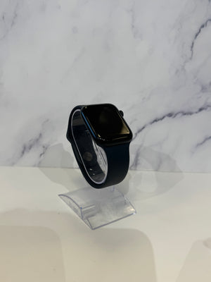 A black smartwatch with a rectangular screen and black sport band, displayed on a clear acrylic stand against a white marble-patterned background.