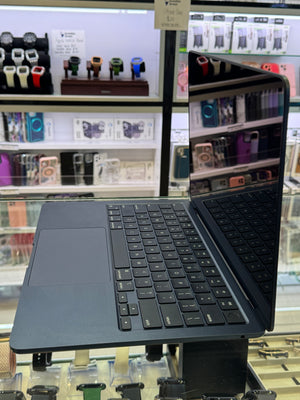 A dark slate blue or gray laptop with a black keyboard and trackpad, shown open on a display stand inside a retail store. Shelves of other electronics and accessories are visible in the background.