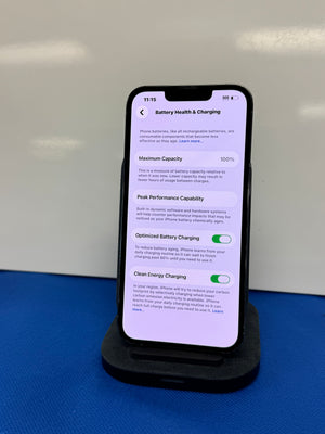 A smartphone displayed upright on a black charging stand against a light gray background, with a blue surface beneath the stand. The phone screen shows battery health and charging settings.