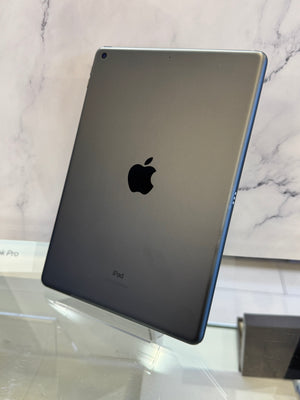 A dark gray Apple iPad Pro shown from the back, resting on a clear glass display stand against a white marble-patterned background.