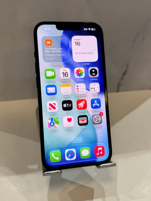 A black Apple iPhone with a lit home screen, displayed upright on a small metal stand on a light beige surface against a neutral gray marble-patterned background.
