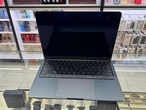 A dark gray Apple MacBook laptop with a black screen and keyboard, sitting on a glass display counter in a retail store.