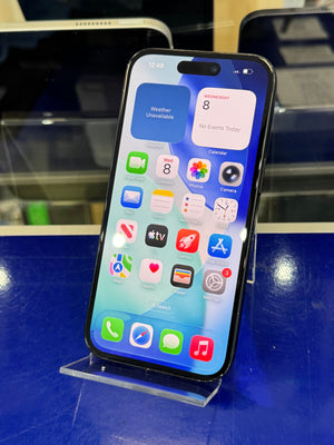 A black Apple iPhone with its screen turned on and displaying the home screen, resting on a clear acrylic display stand against a blue counter.