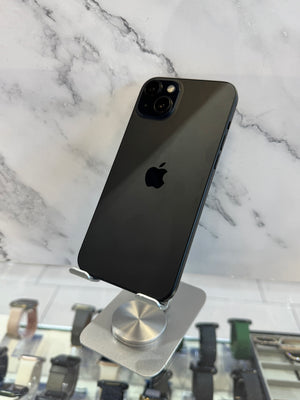 A black Apple iPhone 12 Pro displayed on a silver metal stand, showing its back panel with the Apple logo and triple-camera system, set against a white marble background.