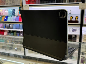 A black iPad case with an integrated keyboard and a subtle embossed Apple logo, shown standing open on a glass display counter in a retail store.