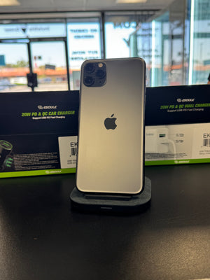 A silver Apple iPhone 12 Pro shown from the back, standing on a small black charging dock on a dark tabletop. Behind it are two black and green Geekle car charger boxes, with a blurred storefront window in the background.