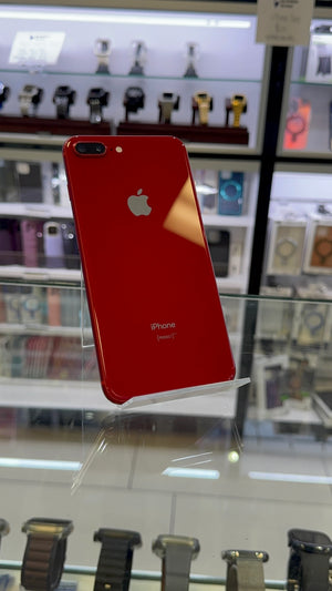 A red Apple iPhone 7 Plus shown from the back, resting on a clear glass display stand inside a retail store with watch displays in the background.