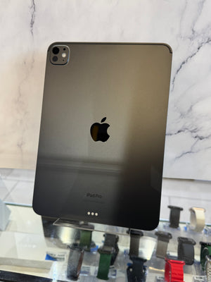 A dark gray Apple iPad Pro shown from the back, resting on a glass display case with other watches in the foreground and a white marble-patterned background.