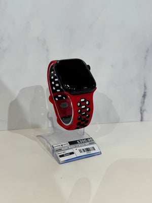 A smartwatch with a black rectangular face and a red perforated sport band, displayed on a clear plastic stand with a price tag in front, against a light marble-patterned background.