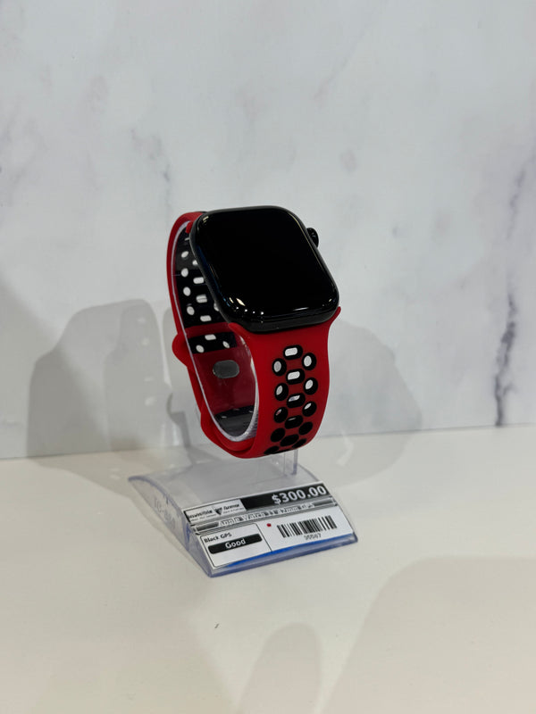 A smartwatch with a black rectangular face and a red perforated sport band, displayed on a clear plastic stand with a price tag in front, against a light marble-patterned background.