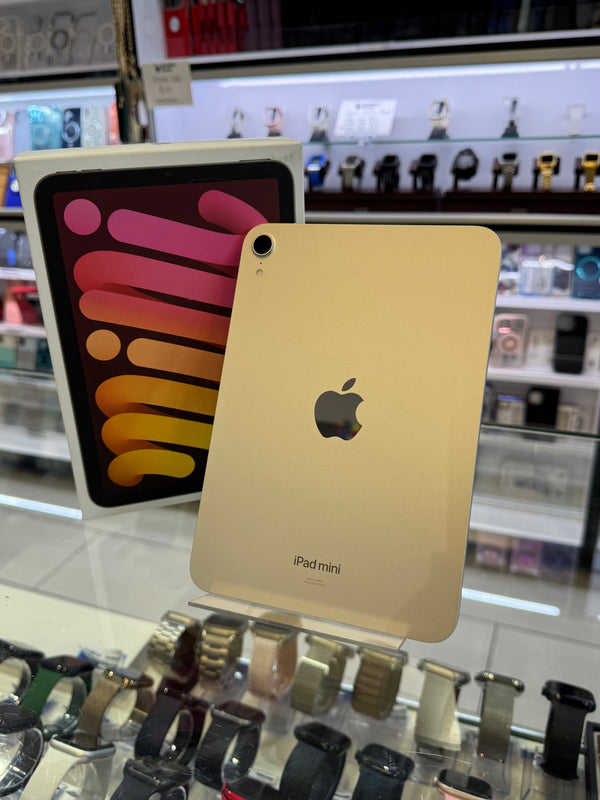 A gold Apple iPad mini standing upright on a display stand in a retail store, with its retail box visible behind it and watch displays on shelves in the background.