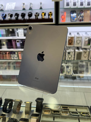 A silver Apple iPad shown from the back, resting on a glass display case inside a retail store with shelves of watches and accessories in the background.