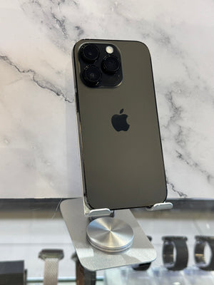 A dark gray Apple iPhone 12 Pro Max standing upright on a silver metal display stand, with a marble-patterned background and watch straps visible in the blurred foreground.