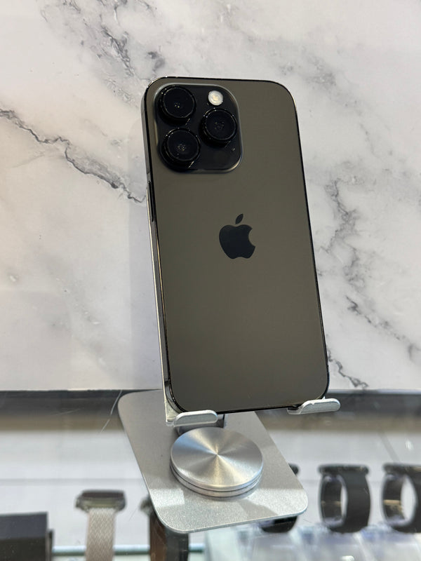 A dark gray Apple iPhone 12 Pro Max standing upright on a silver metal display stand, with a marble-patterned background and watch straps visible in the blurred foreground.