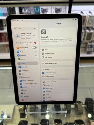 An iPad displaying the Apple Settings app, standing on a glass display stand in a retail store environment.