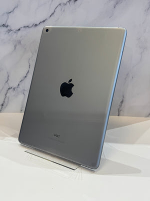 A silver Apple iPad shown from the back, standing on a clear display stand against a white marble-patterned background.