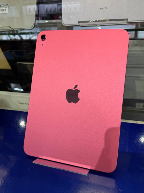 A pink Apple iPad shown from the back, featuring the black Apple logo in the center and a camera lens in the top-left corner, photographed in a retail or showroom setting.