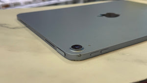 A close-up shot of the back corner of a gray Apple iPad resting on a light beige marble surface. The Apple logo is visible in the upper right, and the camera lens and side buttons are visible on the lower left.