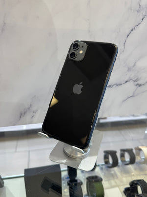 A black Apple iPhone with a glossy glass back is displayed on a silver stand in a retail setting, with a marble-patterned wall in the background.