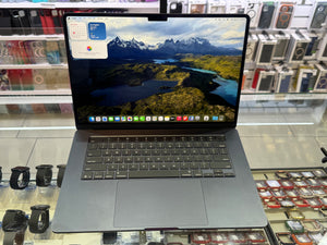 A dark gray Apple MacBook laptop with an open screen displaying a mountain landscape wallpaper, set on a glass display counter in a retail store.