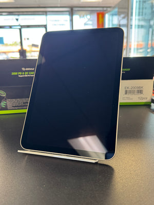 A black tablet with rounded corners displayed upright on a small stand on a dark tabletop, with product packaging boxes visible in the blurred background.
