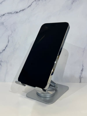 A silver metal adjustable phone stand holding a black smartphone, set against a white marble-patterned background.