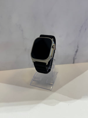 A smartwatch with a black strap and silver-toned case, displayed on a clear acrylic stand against a light gray marble-patterned background.